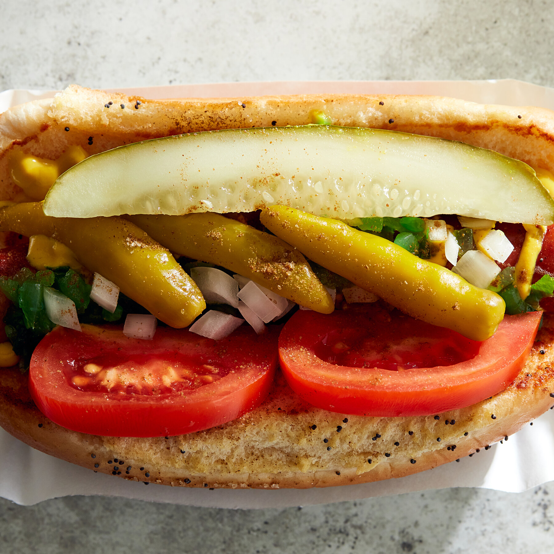 A grilled hotdog sits atop a golden poppyseed bun. This is the Chicago Dog, topped with yellow mustard, green relish, white onion, tomato slices, and yellow sport peppers.