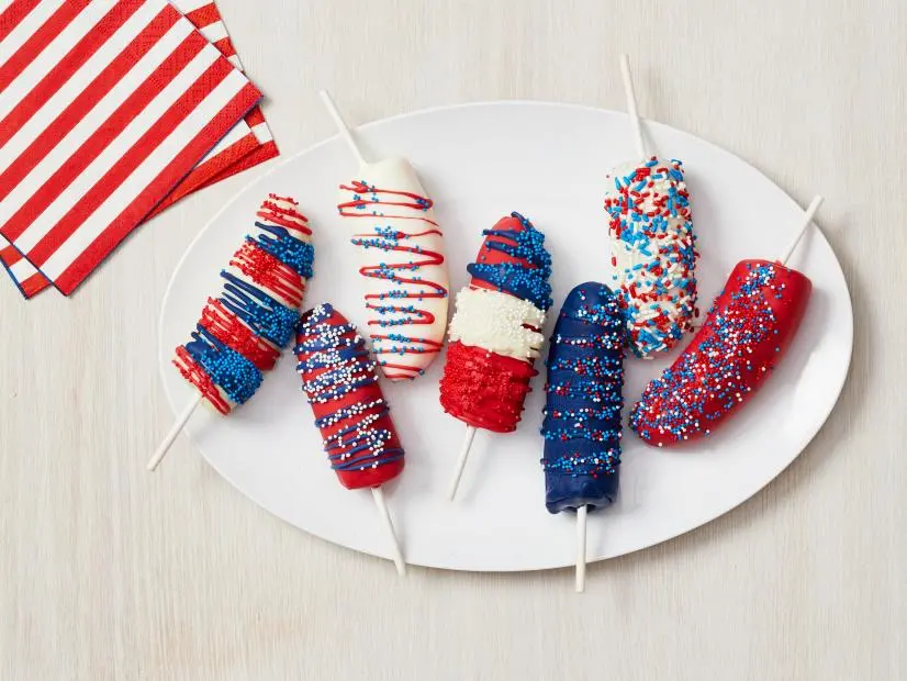 Bananas covered in red, white, and blue frosting with sprinkles and drizzles of different colored chocolate sit on a white oval plate accompanied by red and white horizontal striped napkin.