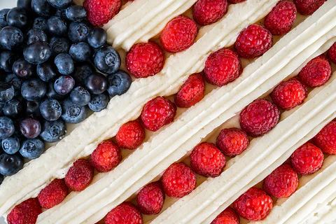 A frosted berry cake is pictured with white cream cheese frosting lines, separated by red raspberry lines representing the stripes of the American Flag, with a blueberry square in the top lefthand corner.